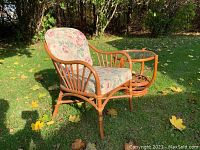 Angle view of the rattan chair with floral cushions and adjacent round rattan table with glass top, placed on grass outdoors with fallen leaves.