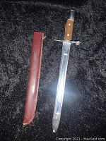 Small sword with wooden handle and accompanying brown leather sheath laid out on black fabric background.