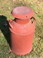 Full view of the vintage metal milk can placed on grass with visible handles and lid.