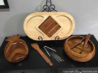 Full view of wood pineapple hostess set showing oval cheese board with dark inset, pineapple shaped bowl, wooden bowl with tongs, wooden fork-knife tongs and metal pieces