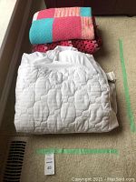 Overall view of folded blankets and quilted mattress cover laid on carpet.