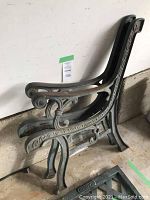 Side view of two cast iron garden bench side frames stacked together showing decorative scroll and leaf design on armrest and legs.
