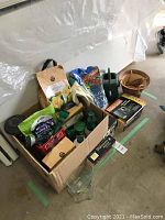 View showing cardboard boxes with assorted garden products including bags of fertilizer, birdseed, gardening gloves, round wooden container, and various tools.