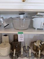 Shelf showing food processor parts including motor base, mixing bowl, four metal blades/discs, and attachments, with salad spinner and rice cooker above.