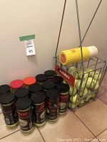 Photo showing a group of sealed Penn tennis ball cans with black lids and red or orange labels next to a metal basket with several loose tennis balls and a yellow canister.