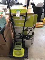 Front view of Sun Joe tiller and cultivator with original box behind, showing green and black color scheme and upright handles.