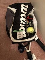 Wilson tennis bag in black and white with Wilson logo, partially open showing tennis balls and rackets handles on carpeted floor