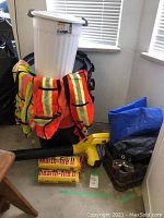 Wide shot showing two orange safety vests, white and black garbage cans, yellow blower, fire logs, and tarps.
