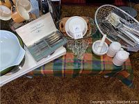 Photo showing green Pyrex bowl, flatware set in original box, marble mortar and pestle, eggs cups, salt and pepper shakers arranged on plaid tablecloth with wire baskets containing utensils and silverware.