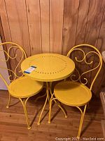 Full view of yellow metal bistro table with two matching chairs against wood paneling.