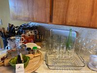 Wide view of kitchen counter with glass wine glasses, beer glasses, cutting boards, wooden utensils, and various glass containers.