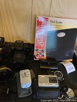 Full lot photo showing Canon EOS film camera, Panasonic Lumix camera, universal travel adapter, compact cassette recorder, and electronic scale in box.