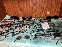 Multiple eyeglasses and sunglasses laid out on a green towel against a wooden background. Various frame styles visible including rectangular, oval, and round.