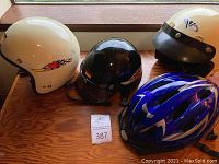 Four helmets on wood surface: one white motorcycle helmet with racing decal, one black motorcycle helmet, one cream-colored motorcycle helmet with studs, one blue bicycle helmet