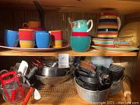 Shelves of kitchen items including colorful ceramic mugs and cups, metal mixing bowls, and KitchenAid mixer accessories.