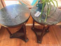 Top view of both round marble topped tables showing green marble surfaces with natural veining and dark cherry wood bases with brass trim and accents, accompanied by a small aloe vera plant.