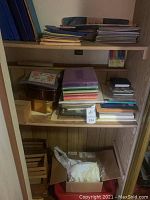 Wide view of shelf with various office and craft supplies including notebooks, paper, paint, and storage boxes.