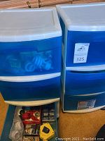 Two plastic storage bins, white body with blue semi-transparent drawers shown full of various items including compact fluorescent light bulbs and small miscellaneous electronics.