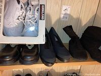 Photo of assorted women's shoes on wooden shelves including white Kirkland sneakers in box, black dress shoes, and black boots.