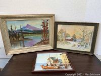 Three framed oil paintings: left a purple mountain lake scene in a light tan frame, center a sailboat painting with a wooden dock in a dark brown frame, right a snow-covered cottage scene in a darker brown frame.