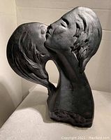 Side view of black stylized statue showing two female faces kissing with detailed hair textures.