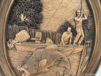 Close-up image of the wood carved plaque showing two men fishing with a large fish hooked, detailed carving with textured trees in background.