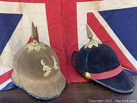 Two Oddfellows regalia helmets, one brown and one dark blue, displayed side by side in front of the Union Jack flag.