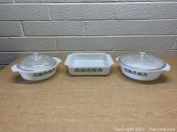 Front view of 3 vintage Anchor Hocking white glass baking dishes with green and yellow floral pattern, two round and one rectangular, all with clear glass lids, on wooden table with brick wall background.