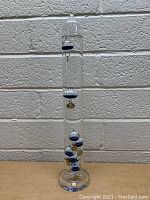 Full view of 17 inch tall glass Galileo thermometer standing on a beige surface against a gray brick wall. Shows entire thermometer with floating spheres and tags inside glass tube.