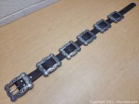 Full view of brown leather belt with seven detailed silver buckles laid out on a wood surface.