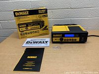 DeWalt battery charger and maintainer with box and manuals on table in front of brick wall, unit shown turned off.