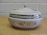 Front view of round handmade porcelain bowl with lid, showing gold leaf accents, white flowers, and bead detailing.