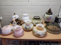 Photo showing assortment of vintage hand-painted glass lamp shades and hanging lamps on a table in front of a white brick wall.