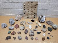 Photo showing a large assortment of rough rocks and geodes of various sizes and colors displayed on a table with a labeled mineral specimen board in the background.