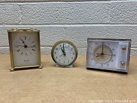 Three vintage clocks shown from front and angled side views on wooden surface against cinderblock wall.