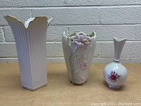 Three Lenox vases arranged on a shelf, showing distinct shapes and designs including a tall white fluted vase, a sculpted floral relief vase, and a small round vase with floral decal.