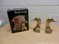 Photo showing the pair of brass duck head bookends and their original packaging box on a table.