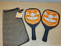 Pair of new orange and black pickleball paddles with white graphics and text that reads 'Pickleball' on the paddle face, shown along with a black mesh carrying bag with drawstring and brand tag attached.