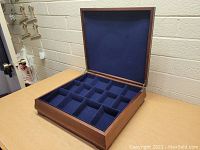 View of wooden display case with open lid showing 15 blue velvet-lined compartments inside.