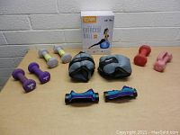 Photo shows all items on a beige table against a white brick wall, including various dumbbells, ankle weights, hand grips, and a boxed exercise ball.
