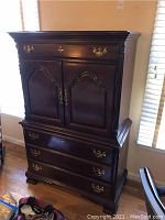 Front view of dark wood armoire chest with brass hardware, ornate carved panels, three large drawers below double cabinet doors, and top smaller drawer.