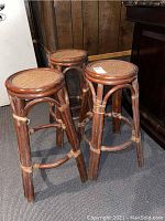 Three wooden bar stools with woven cane seats grouped closely together, showing the round seats and structural details including binding accents and scratches.