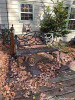 Full view of outdoor cast iron bench placed on outdoor decking surrounded by dry leaves. Shows overall structure and decorative backrest.
