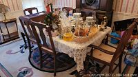 View of the dining room table with lace tablecloth and six chairs surrounding it. Table holds various decorative items not included in lot.