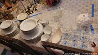View of Noritake china dinnerware stacked including plates, bowls, saucers, Lenox covered bowl, and variety of stems and glasses, candle holders on floral tablecloth.