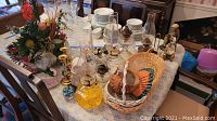 Photo shows vintage oil lamps, candle holders, woven baskets with ceramic poultry figurines, Cynthia Rowley tea cup and saucer, faux flower arrangement and various small decorative figurines on a table.
