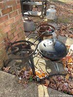 Two rusty metal propane burners beside a black Weber kettle grill with lid outdoors on a leaf-strewn surface.