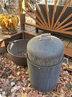 Two outdoor cooking items on ground covered with autumn leaves: a vertical black metal smoker with dome lid and side handle, and a large cast iron cauldron bowl behind it.