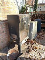 Full view of the metal smoker placed outdoors showing front door, legs, and side handle with rust and grime on surface.