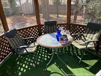 Photo of the outdoor wicker round table with glass top surrounded by three padded folding chairs placed on outdoor turf in a screened porch area.